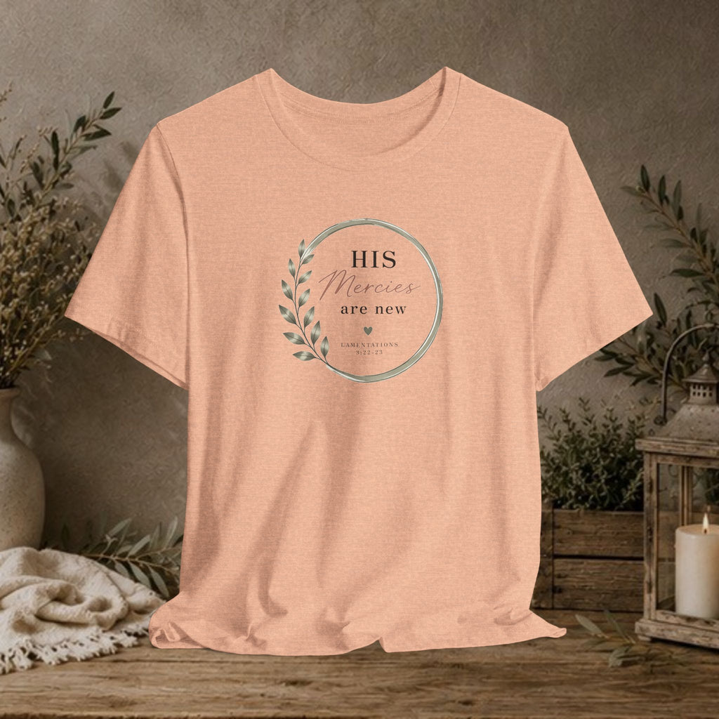 A peach-colored short-sleeve t-shirt with a circular graphic on the chest reading 'HIS Mercies are new' with a leaf design, displayed on a rustic wooden table with a neutral background and decorative plants.