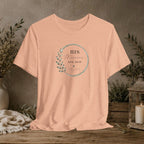 A peach-colored short-sleeve t-shirt with a circular graphic on the chest reading 'HIS Mercies are new' with a leaf design, displayed on a rustic wooden table with a neutral background and decorative plants.