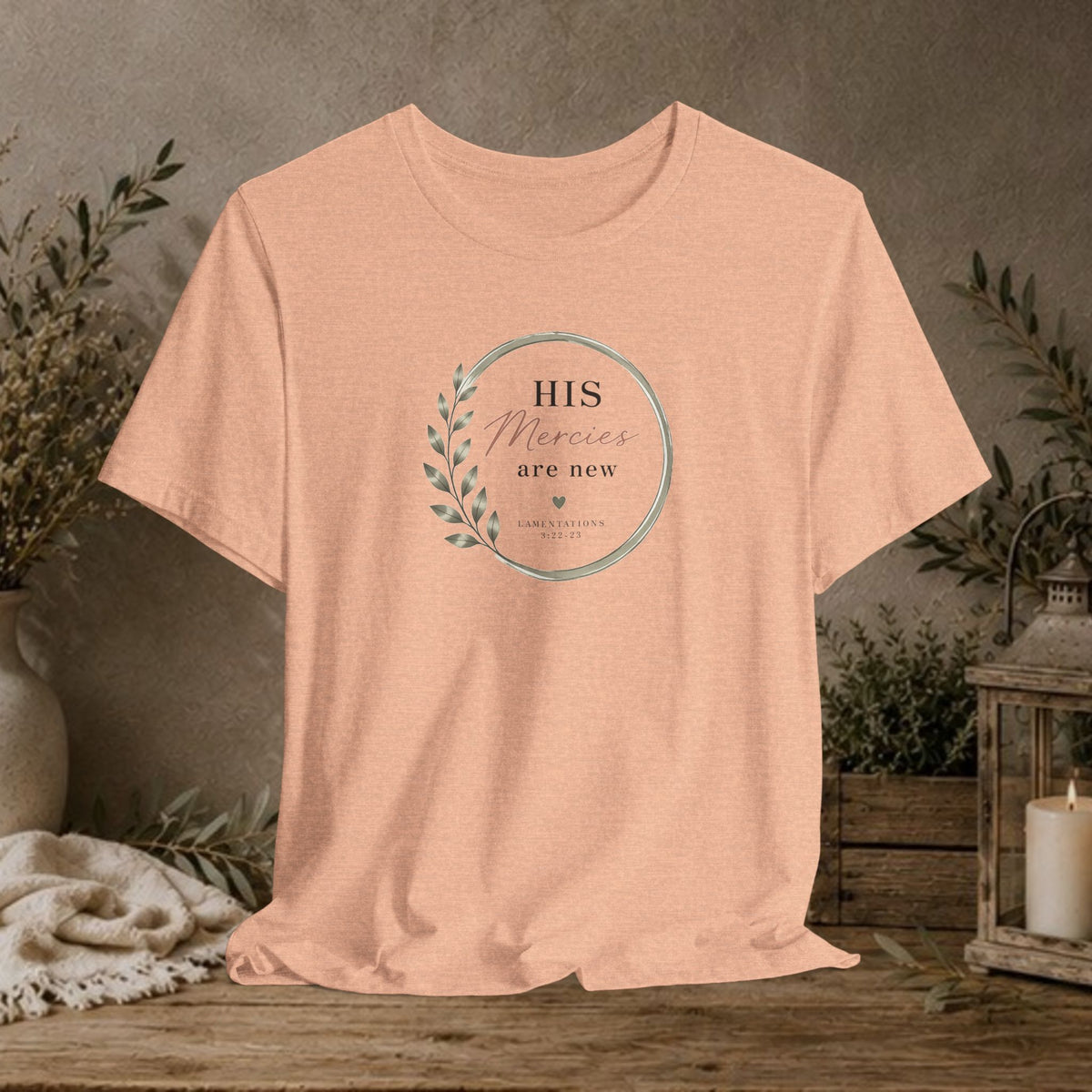 A peach-colored short-sleeve t-shirt with a circular graphic on the chest reading 'HIS Mercies are new' with a leaf design, displayed on a rustic wooden table with a neutral background and decorative plants.